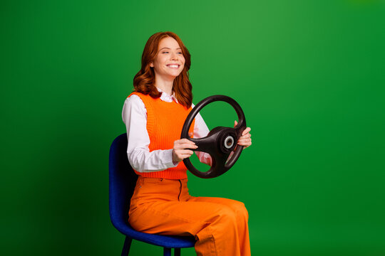 Smiling woman holding a car steering wheel while pretending to drive against a green studio background wearing a bright orange outfit