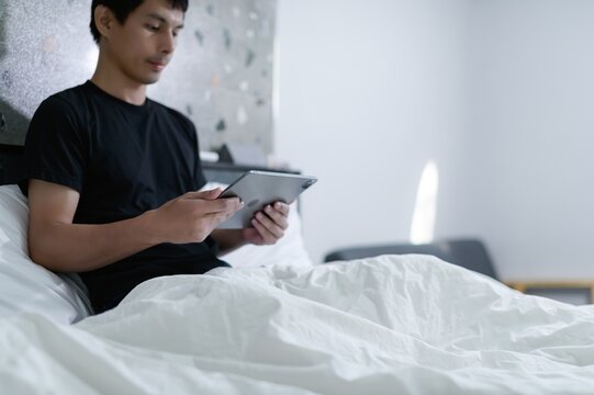 Young man using tablet while sitting on bed in cozy bedroom setting