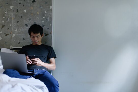 Young Man Using Smartphone While Sitting Next to Laptop on Bed