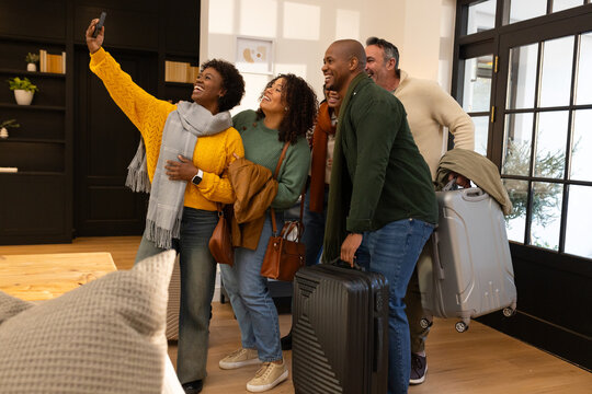 Diverse friends posing for selfie in home entryway, holding smartphone and two suitcases
