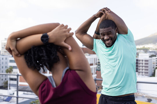 African American friends stretching on rooftop by metal railing, wearing maroon tank and smartwatch