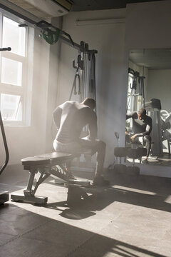 African man in compression gear sitting on bench in gym by cable machine holding dumbbells