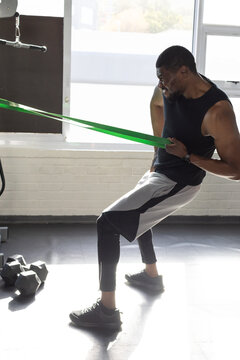Mid-adult African-American man in black top pulling green band in gym with hex dumbbells