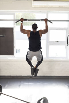 African American man performing pull-up on metal bar at gym by windows wearing black tank