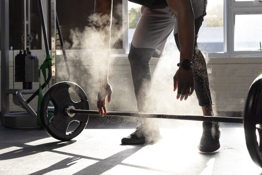African American man chalking hands and preparing heavy barbell at gym near windows