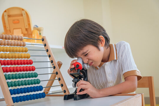 Generation Alpha boy peers through toy microscope beside colourful wooden abacus, showing scientific and mathematic interested and cognitive development during kindergarten STEM activity in young age