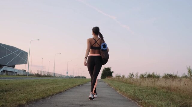 Rear view of young woman walking on paved path beside road at dusk while carrying blue rolled yoga mat tucked into textured woven shoulder bag, with woman, walking, yoga, mat detail