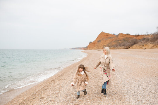 Happy child girl running with her mother at beach wearing beige jackets at sandy beach together. Autumn season.