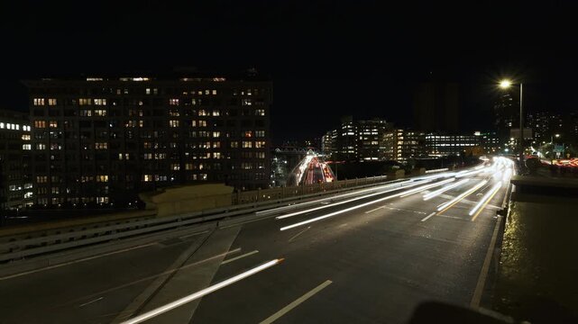 Dense parallel light trails from headlights and taillights flow along curved multilane elevated highway beside large illuminated midrise building, overlapping white streaks concentrated in right