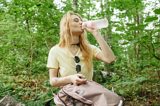 Young adult Caucasian woman drinking water from bottle during forest hike, staying hydrated. Image suitable for outdoor recreation, eco tourism and healthy lifestyle marketing campaigns