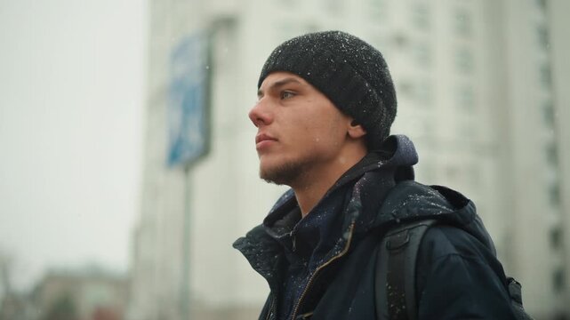 Close up of man wearing dark winter clothing and backpack standing outdoors in falling snow with tall building visible in background. Jacket beanie covered by wetness from precipitation
