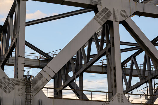 Intricate steel truss framework with riveted connection plates, metal ladders and railings against pale blue sky with scattered soft clouds.