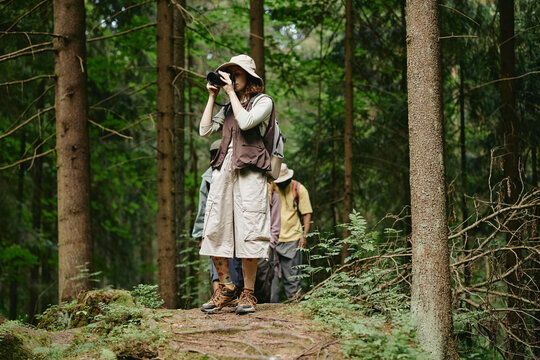 Diverse young adult friends hiking in forest while Caucasian woman takes photo with camera. Image ideal for eco tourism campaigns and outdoor adventure gear marketing, eye level shot