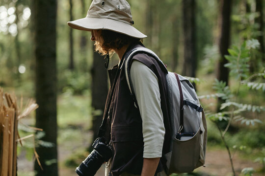 Side view of young Caucasian woman hiking in forest with backpack and camera. Image represents outdoor recreation and is suitable for travel and adventure tourism marketing