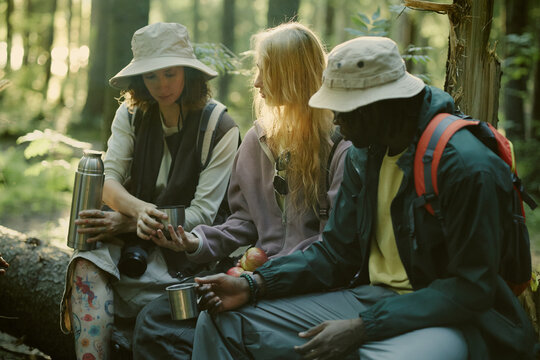 Diverse young adult friends sitting on log, drinking tea from thermos during forest hike. Image suitable for outdoor recreation gear marketing and eco tourism campaigns