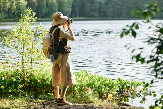 Young adult Caucasian woman hiking in forest, standing near lake and taking photos with camera. Image represents outdoor recreation, suitable for travel and adventure marketing campaigns