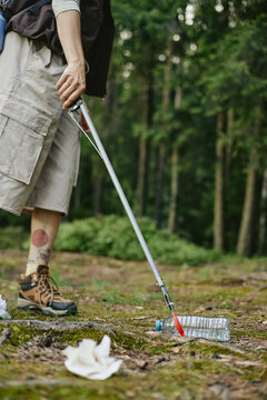 Vertical shot of young adult Caucasian man using trash grabber tool to pick up plastic bottle in forest, promoting environmental cleanup campaigns and eco friendly tourism