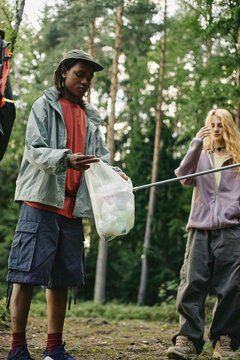 Vertical image of diverse young adults collecting plastic waste in forest, promoting environmental conservation and eco friendly community initiatives