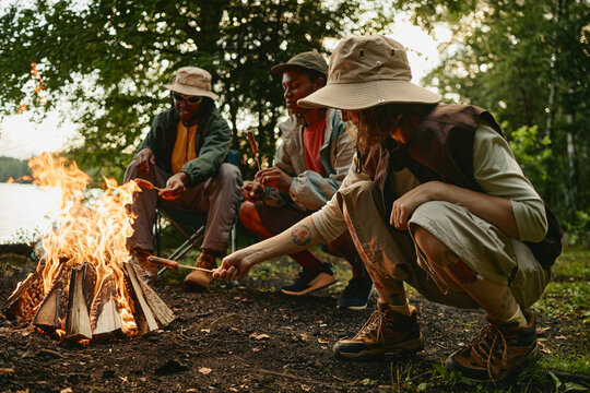 Diverse young adult friends roasting sausages over campfire in forest during hiking trip. Image represents outdoor recreation and is suitable for travel and adventure marketing