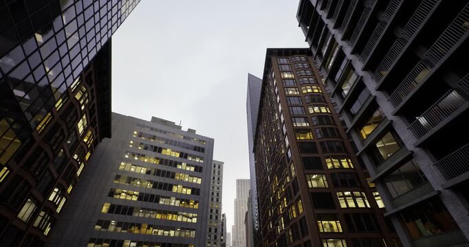 Illuminated office towers at dusk framing tight urban canyon with glass facades reflecting warm interior lights, corporate headquarters rising toward overcast