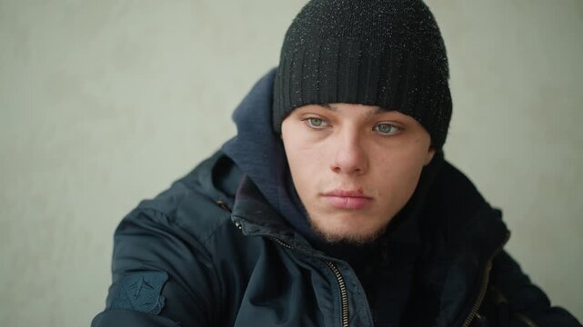Close up of young man wearing winter clothing including black jacket and knit hat seated against plain background. Jacket features visible patch on sleeve subject maintains neutral