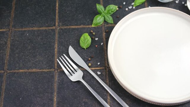 White plate with fork and knife surrounded by basil leaves and peppercorns on dark tiled background