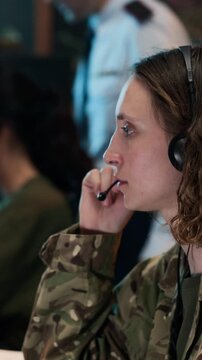 Young adult Caucasian female soldier wearing headset and typing on keyboard in military command center. Footage ideal for defense industry and national security videos, vertical