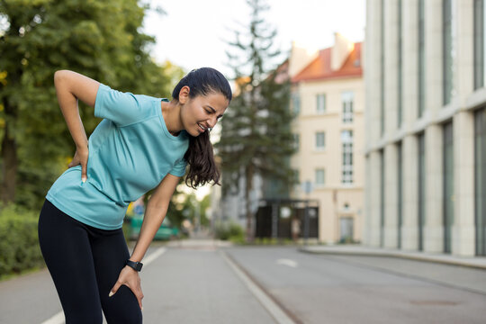 Woman suffering from acute lower back pain and knee discomfort while exercising on a city street, indicating a sports injury or physical strain during her outdoor fitness routine