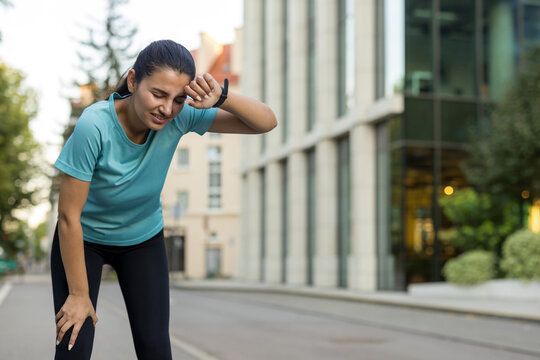 Exhausted woman jogger resting after intense running, wiping sweat from her forehead, feeling tired and fatigued in a city environment during her fitness exercise