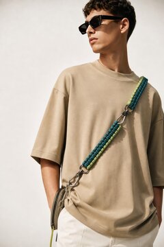Young man in casual streetwear posing confidently with colorful braided shoulder strap and sunglasses, modern urban style, neutral background, lifestyle fashion concept