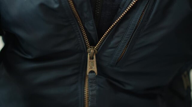 Close up of person wearing dark blue jacket with zipper partially open revealing part of inner garment. focus remains on texture and details of fabric and brass colored zipper. Soft lighting
