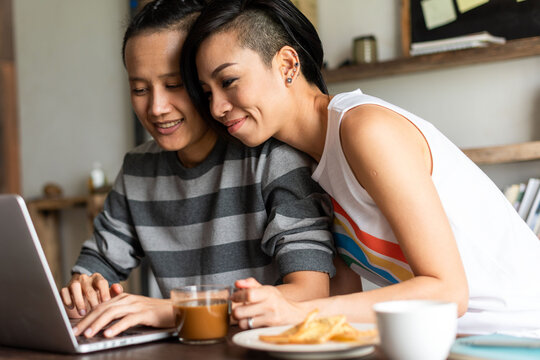 Two people smiling at a laptop, enjoying coffee and snacks together.