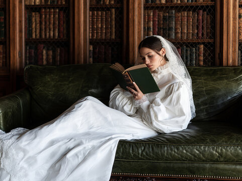 Elegant bride in Victorian lace gown reading old hardcover book on green leather sofa in library
