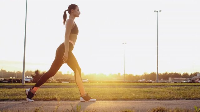 Young woman in athletic wear holds forward lunge on grassy roadside at sunset, right knee lowered near ground and left foot planted with heel contact while arms extend straight ahead