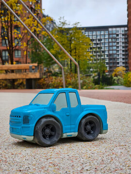 Blue plastic toy truck sits on gravel path. Background features urban buildings and autumn trees. Soft focus adds depth to the scene. Bright blue color draws immediate attention