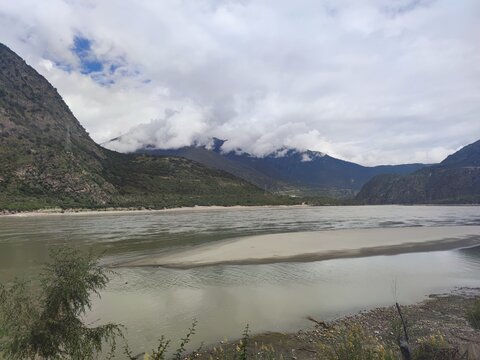A wide braided stretch of the Yarlung Tsangpo river with sandbars and forested mountain slopes under dramatic clouds near Pai Town in the Grand Canyon area, Nyingchi, Tibet, China