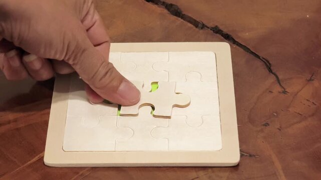 Close-up shot of a hand placing the final missing wooden jigsaw puzzle piece into place to complete the wooden puzzle on a wooden surface. Concept of problem solving, success, achievement, completion,