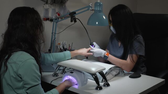 Manicure technician spot cures single nail while client dries other hand fully under ultraviolet lamp during salon procedure