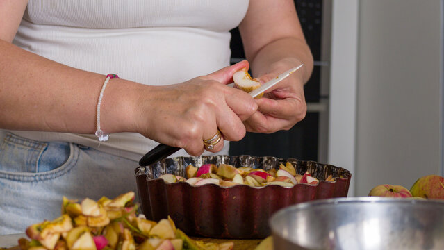Close-up of person's hands skillfully using small paring knife to slice fresh apples into rustic metal baking pan while preparing homemade apple pie or fruit dessert at home