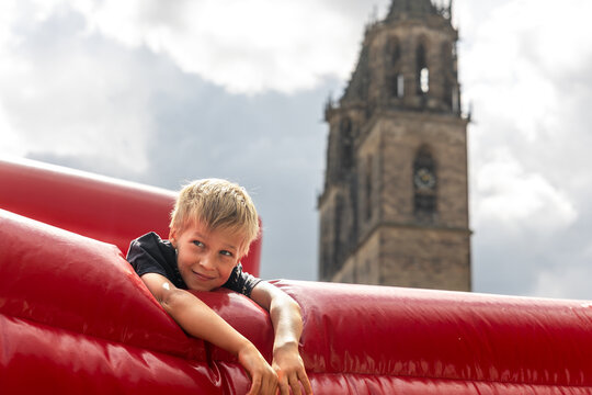 Smiling boy relaxes on bright red inflatable attraction in Magdeburg, with historic Dom cathedral tower behind him. Sunny urban scene combines childhood fun, landmark architecture, leisure and travel
