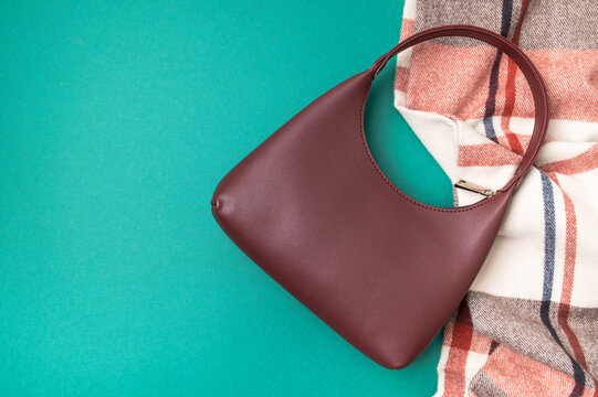 Burgundy leather handbag and plaid scarf rest on green background with copy space. Stylish purse and warm textile create seasonal fashion composition for retail advertising and autumn campaigns.