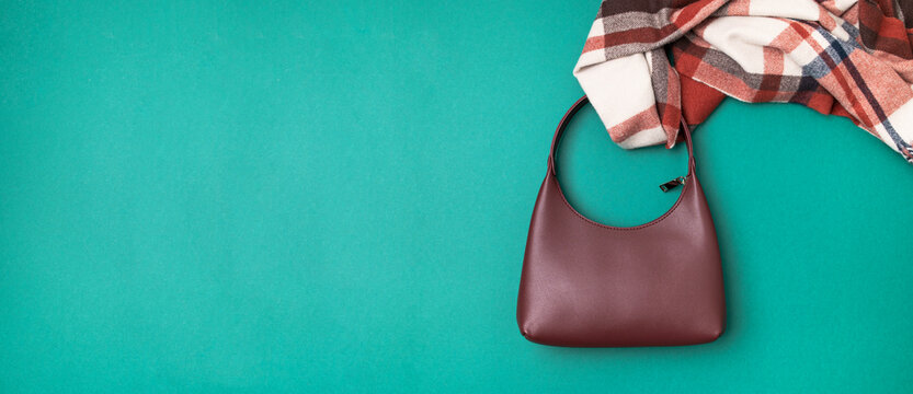 Burgundy leather female baguette bag and checkered warm scarf on green background top view. Fashion and style concept, womens accessories. Autumn wardrobe items. Space for text.