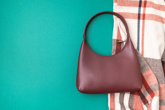 Burgundy leather baguette bag rests beside plaid scarf on green background, presenting stylish fashion accessories, autumn wardrobe details and copy space for retail marketing campaigns.