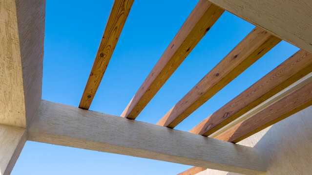 View from below of modern pergola with textured concrete supports and parallel wooden beams against clear blue sky on sunny day