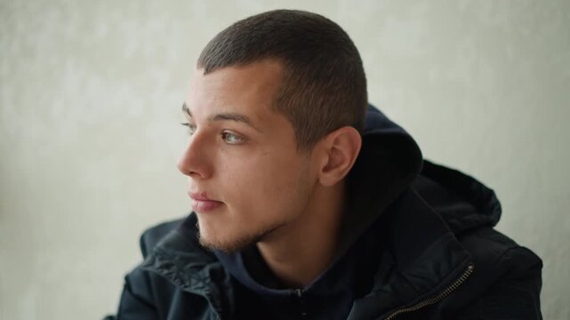 Close up of young man adjusting black over ear headphones while wearing dark jacket and hoodie seated indoors with plain light colored wall background. Subject maintains calm expression