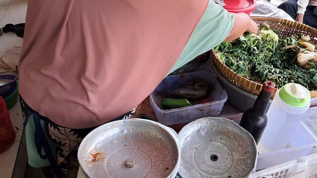 Asian street food vendor cutting ingredients into traditional stone mortar to prepare Indonesian gado gado salad with spicy peanut sauce.