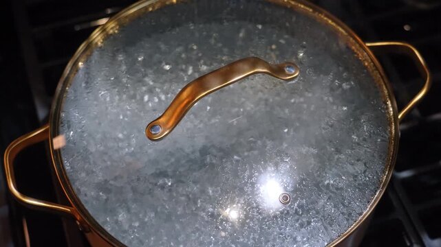 Metal cooking pot with glass lid on stove. Steam condensation covers cover while water boils inside container.