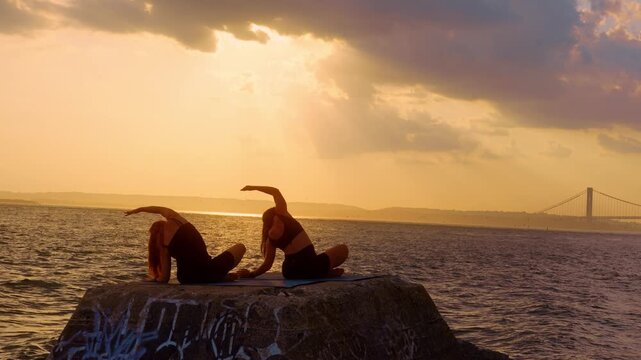 Seated side bend stretch practice on pier, New York, USA. Two female silhouettes stretching upper body against golden sky and suspension bridge backdrop.
