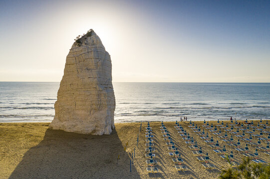 Aerial view of the iconic Pizzomunno limestone monolith standing on a sandy beach with rows of blue and white umbrellas along the Adriatic Sea in Apulia, Italy.