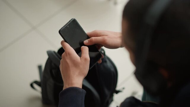 Close up of person holding and interacting with smartphone focusing on hands tapping screen. black backpack is visible in background on tiled floor. Soft lighting creates natural indoor
